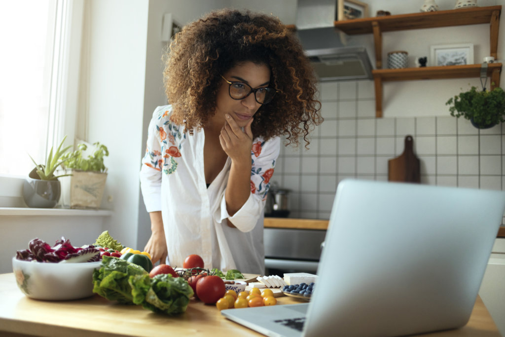 Mulher debruçada no balcão usando o laptop e cercada de alimentos. Ela tem o semblante concentrado, de quem quer aprender a cozinhar