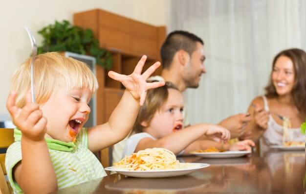 Família de quatro pessoas sentadas à mesa. Eles estão comendo; No primeiro plano, podemos ver um garoto com o rosto sujo e segurando um garfo. Na sua frente está um prato com macarrão, um dos pratos sugeridos para o almoço gostoso e fácil